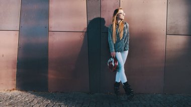 Bina duvarına yaslanmış paten ve kasklı, modaya uygun beyaz bir kadın. Şehir hayatı