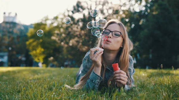 Parktaki çimenlerde uzanırken sabun köpüğü üfleyen mutlu beyaz kadın.