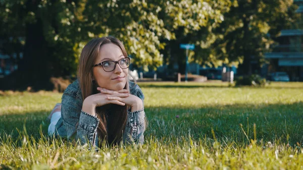 Jonge Kaukasische brunette met bril liggend op het gras in het park en denkend. Ontspanning in de natuur — Stockfoto