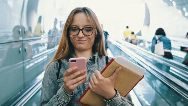 Портрет студента, использующего смартфон и держащего книги и планшет на движущейся лестнице — стоковое фото