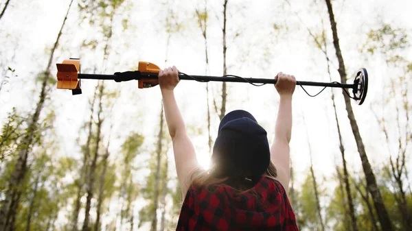 Treasure hunter, young woman holding metal detector in the air in the forest