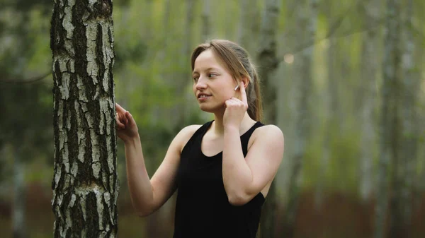 Doğada beyaz bir kadın var. Kablosuz kulaklıklardan müzik dinlemek ve ağaç kabuğuna dokunmak. Yavaş çekim. — Stok fotoğraf