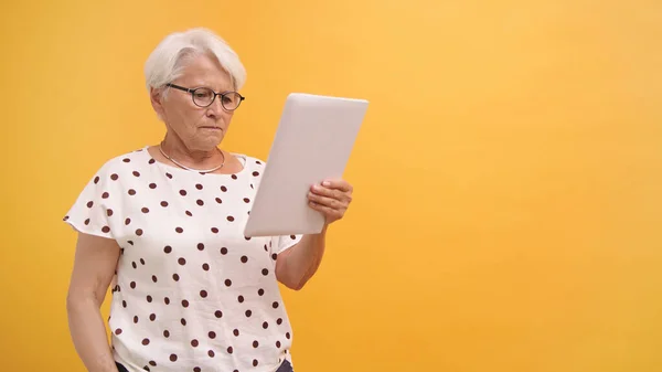 Una señora mayor confusa sosteniendo una tableta de regalo buscando ayuda. Aislado sobre fondo naranja — Foto de Stock