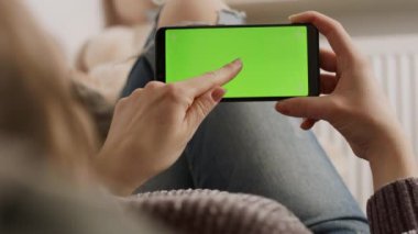 Kanepede dinlenirken akıllı telefonun yeşil ekranına dokunan genç kadın.