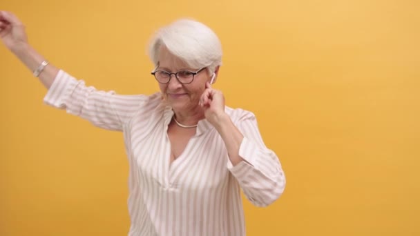 Divertida anciana feliz bailando con auriculares inalámbricos, aislada sobre fondo naranja — Vídeos de Stock