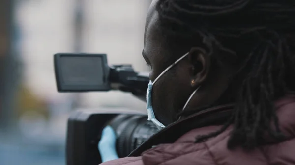 Yakından, tıbbi maskeli Afrikalı siyahi videograf video kameradaki görüntü bulucudan bakıyor. — Stok fotoğraf