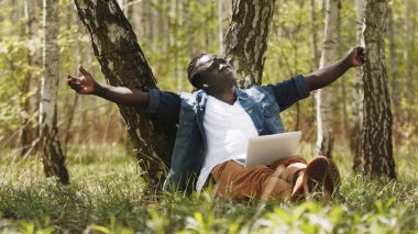 Uzaktan çalışma konsepti. Doğada dizüstü bilgisayar kullanan elleri açık Afrikalı bir adam.