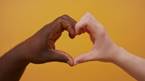 Manos blancas y negras formando forma de corazón juntas aisladas sobre el fondo naranja. De cerca. — Vídeos de Stock