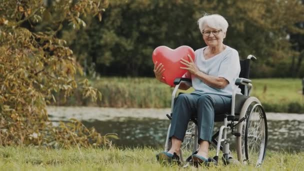 Старшая женщина в инвалидной коляске возле реки держит шарик в форме сердца — стоковое видео