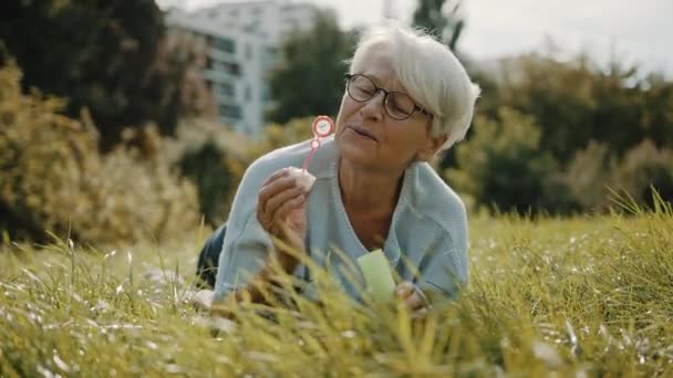 Rentnerin genießt Freiheit. Kindliche Oma pustet Seifenblasen im Park — Stockvideo