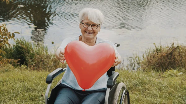 Старшая женщина держит шарик в форме сердца у реки. Любовь и забота — стоковое фото