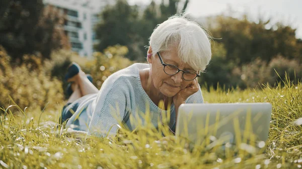Anciano jubilado mujer usando el ordenador portátil en la naturaleza mientras está acostado en la hierba — Foto de Stock