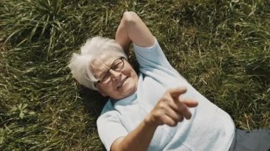 Yaşlı kadın çimenlerde dinleniyor ve parmaklarıyla havaya kalp şekilleri yazıyor.