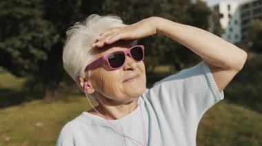 Parkta pembe güneş gözlüklü yaşlı kadın mesafeye bakıyor.