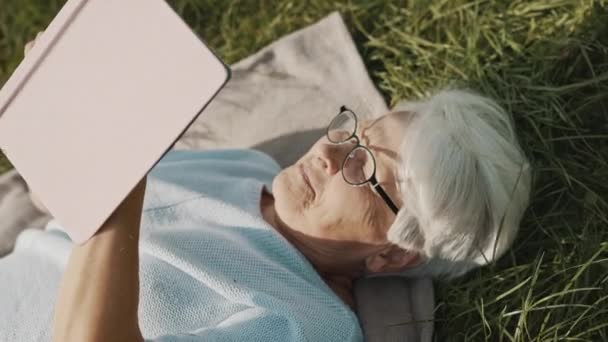 Gepensioneerde grijze harige vrouw die een boek leest terwijl ze rust op het gras in de natuur — Stockvideo