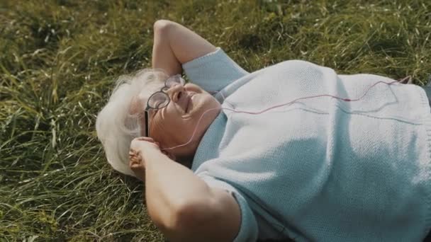 老婦人はヘッドフォンを草の上に寝そべって音楽を聴き — ストック動画