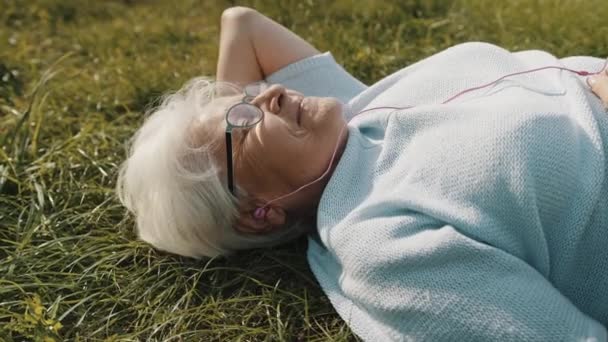 Ältere grauhaarige Frau mit Kopfhörern auf dem Gras liegend und Musik hörend — Stockvideo