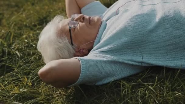 Cool mormor, kvinna med hörlurar liggande på gräset och lyssna på musik — Stockvideo