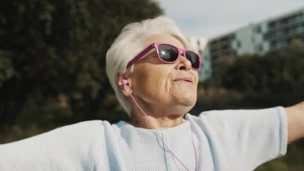 Cool grand-mère avec des lunettes de soleil roses et des écouteurs qui s'étirent dans le parc. Loisirs dans la vieillesse — Video