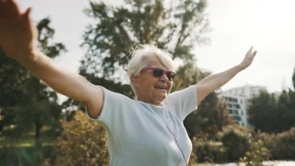멋진 할머니가 분홍색 선글라스를 끼고 공원을 돌아다니며 음악을 들으면서 — 비디오