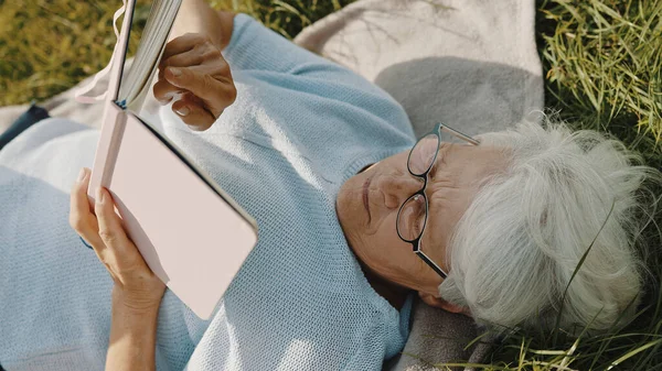 Mujer de pelo gris senior leyendo un libro mientras descansa sobre la hierba en la naturaleza — Foto de Stock