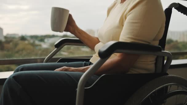 Пожилая женщина в инвалидной коляске на террасе пьет чашку чая или кофе. Без лица — стоковое видео