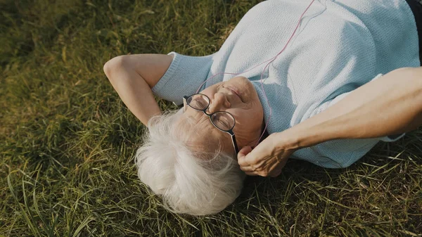 헤드폰을 들고 풀밭에 누워 음악을 듣고 있는 나이든 회색 머리 여자. — 스톡 사진