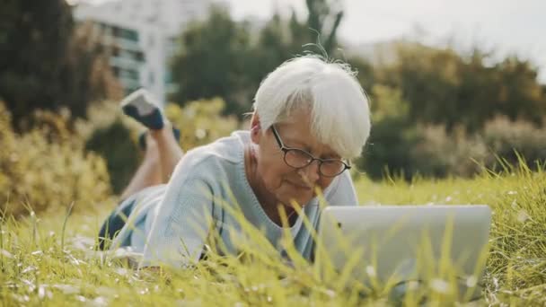 Senior gepensioneerde vrouw met behulp van laptop in de natuur terwijl liggend op het gras — Stockvideo