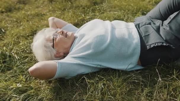 Ωραία γιαγιά ξαπλωμένη στο γρασίδι με τα χέρια κάτω από το κεφάλι της. Ανάπαυση στη φύση — Αρχείο Βίντεο