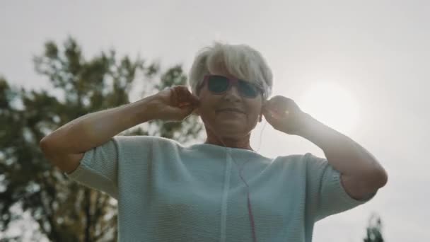 Parkta dans eden pembe güneş gözlüklü ve kulaklıklı havalı bir büyükanne. Yaşlılıkta eğlence. — Stok video
