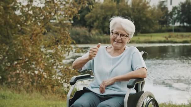 Senior senhora de cabelos grisalhos na cadeira de rodas perto do rio mostrando polegares para cima — Vídeo de Stock