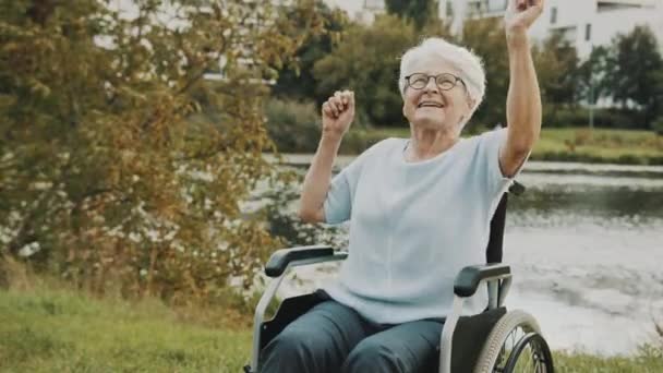 Старшая женщина танцует с руками в инвалидной коляске у реки осенним днем — стоковое видео