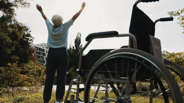 Концепция восстановления. Старуха с руками в воздухе в парке перед инвалидной коляской — стоковое фото