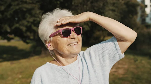 Starsza kobieta w różowych okularach przeciwsłonecznych w parku patrząc na odległość — Zdjęcie stockowe