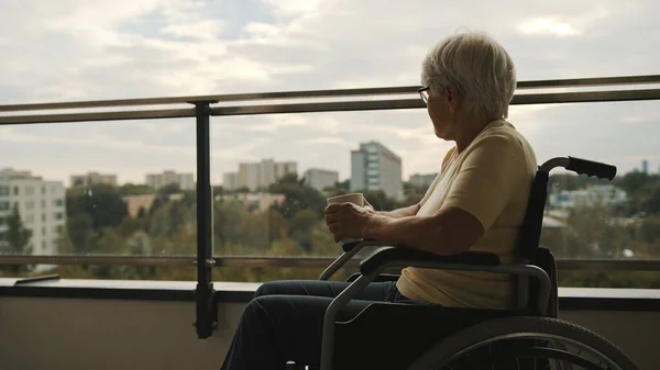 Пожилая женщина-инвалид в инвалидной коляске на террасе пьет чашку чая или кофе. — стоковое фото