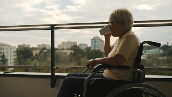 Женщина с седыми волосами в очках пьет кофе в инвалидной коляске на балконе — стоковое фото