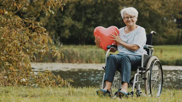Старшая женщина в инвалидной коляске возле реки держит шарик в форме сердца — стоковое фото