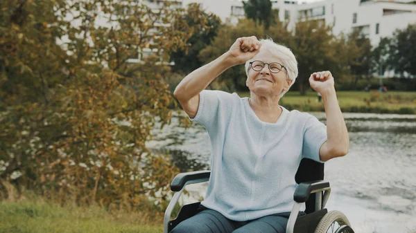 Szczęśliwa staruszka, babcia, tańcząca rękami na wózku inwalidzkim nad rzeką — Zdjęcie stockowe