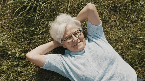 노인 여성, 연금 수급자는 머리 밑으로 손을 아래로 하고 풀 위에 누워 머리 위로 가까이 촬영 — 스톡 사진