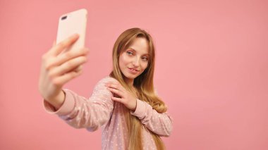 Pijamalı genç kız akıllı telefonu kullanarak selfie çekiyor. Pembe arka planda izole edilmiş