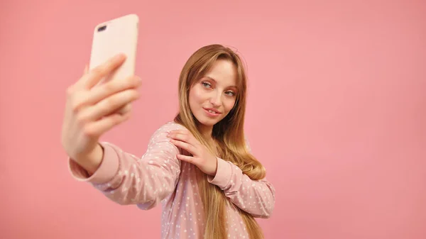 Девочка-подросток в пижаме делает селфи с помощью смартфона. Изолированный на розовом фоне — стоковое фото