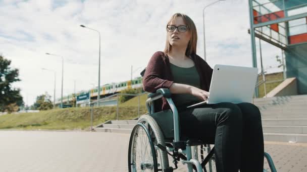 Młoda, biała kobieta na wózku inwalidzkim, używająca laptopa. Przejeżdżający pociąg w tle — Wideo stockowe