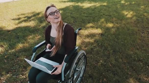 Mujer joven y feliz en silla de ruedas con laptop y smartphone mirando hacia el cielo. — Vídeos de Stock