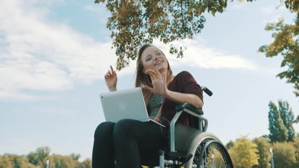 Mujer caucásica feliz en la silla de ruedas escuchando la música en el portátil bajo el árbol. Ángulo bajo — Vídeos de Stock