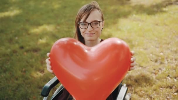 Schöne junge Frau im Rollstuhl hält herzförmigen Ballon im Park — Stockvideo