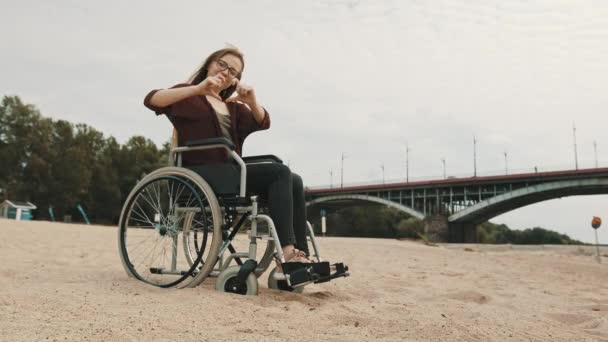 Apoyo a personas con discapacidad. Mujer joven en silla de ruedas en la costa del río de arena mostrando el corazón con las manos — Vídeo de stock