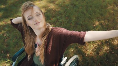 Tekerlekli sandalyedeki genç, engelli beyaz kadın akıllı telefondan müzik eşliğinde parkta sonbahar gününün tadını çıkarıyor.