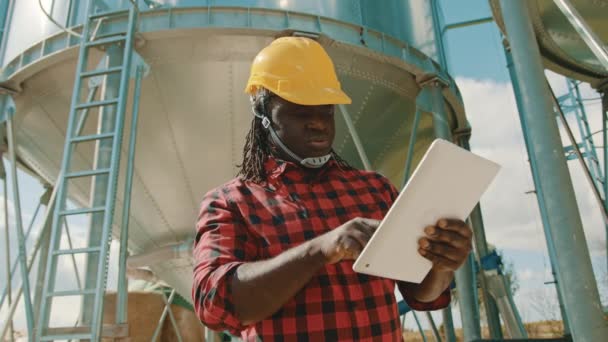 Jonge knappe Afrikaanse man, ingenieur met veiligheidshelm met behulp van tablet in de voorkant van silo-systeem — Stockvideo