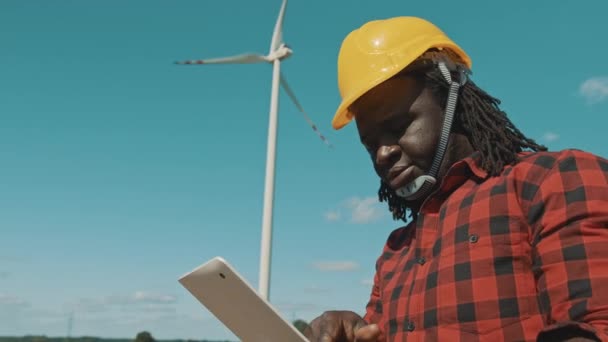 Volwassen of volwassen professionele Afrikaanse man in harde hoed met moderne digitale tablet tegen windmolen maken van inspecties van objecten — Stockvideo