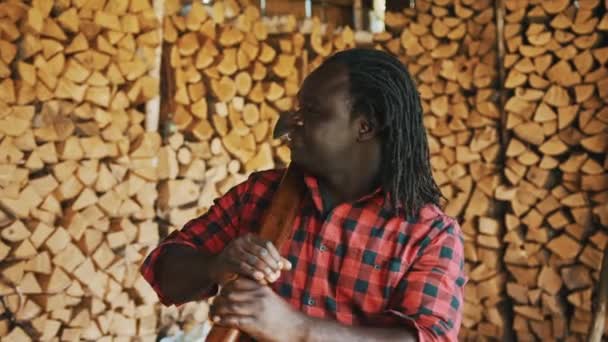 Африканський чоловік з сокирою стоїть перед стосом вирубаних дерев на зиму. — стокове відео